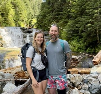 Smiling couple in hiking clothes standing on rocks in front of a forest waterfall